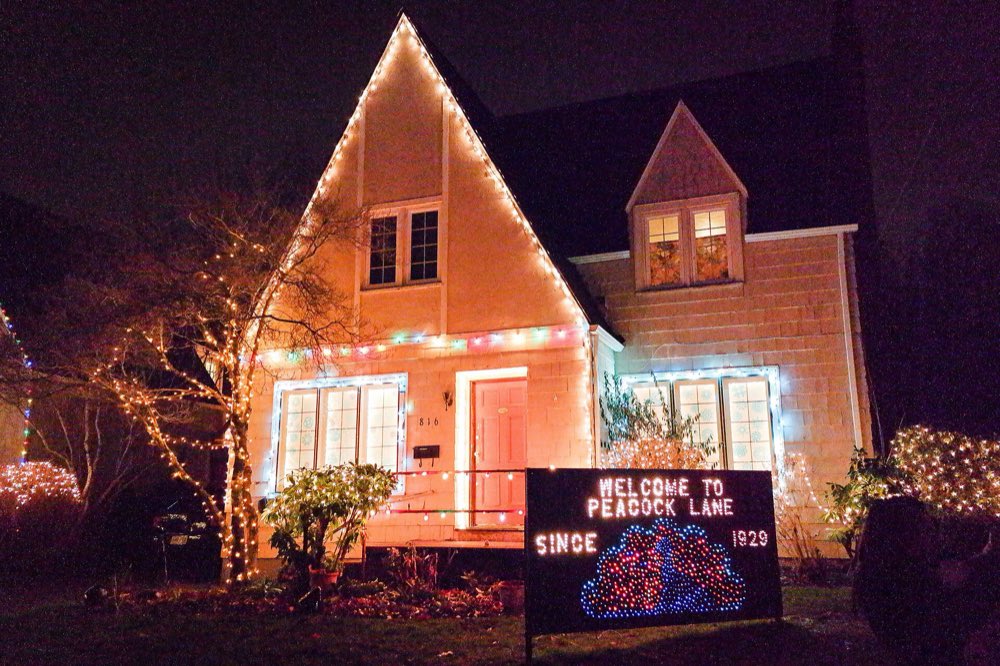 A peaked-roof house on Peacock Lane outlined in warm Christmas lights at night, with a lit sign welcoming visitors to the street