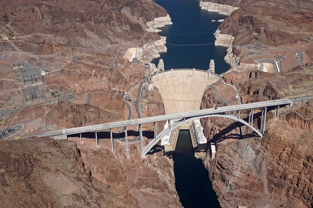 Aerial view of Hoover Dam's pale concrete curve wedged in a red-brown desert canyon, with the arched Memorial Bridge crossing in front and the blue Colorado River below
