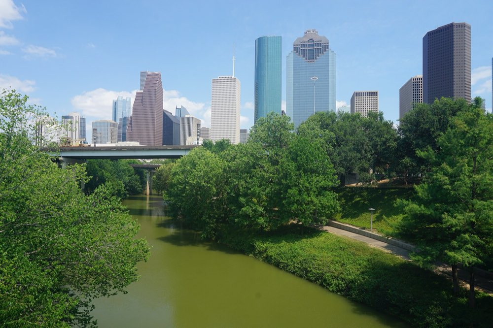 Buffalo Bayou curving through thick green trees and a paved trail in Buffalo Bayou Park, with the glass towers of downtown Houston rising behind under a blue sky