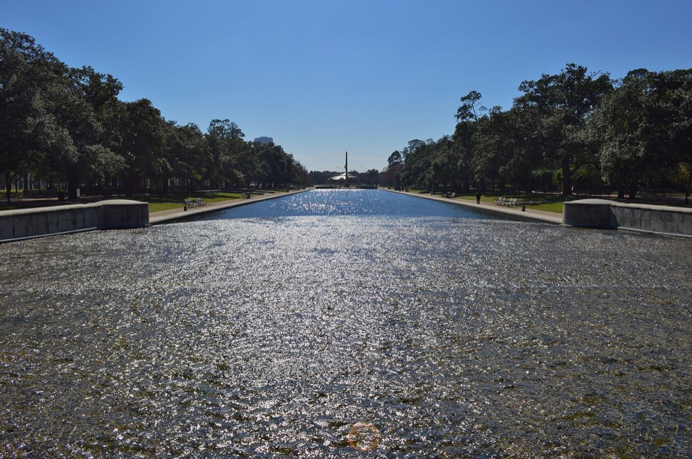 The long Hermann Park reflection pool glittering in the sun between rows of live oaks, with the Pioneer Memorial obelisk at the far end