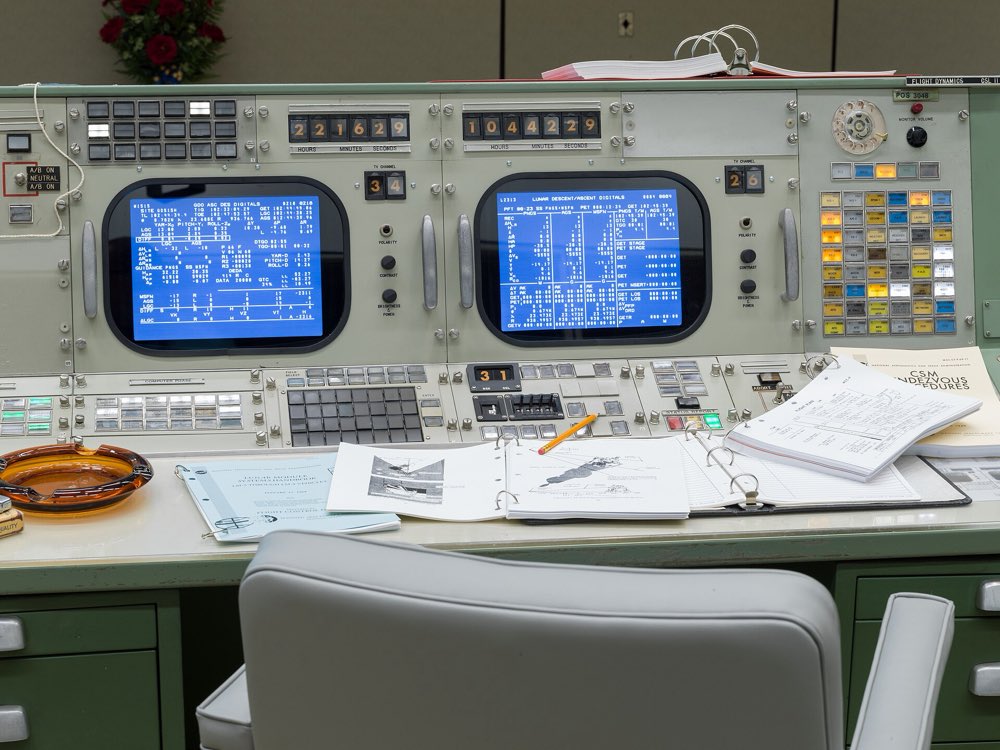 Restored Apollo-era Mission Control consoles with two glowing blue data screens, rows of switches, open flight binders and an empty gray chair