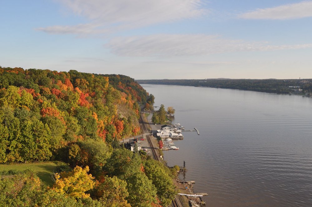 The Hudson River seen from high on the Walkway, with a wooded bluff in orange, red and green fall color dropping to a railroad line and small marina along the west bank