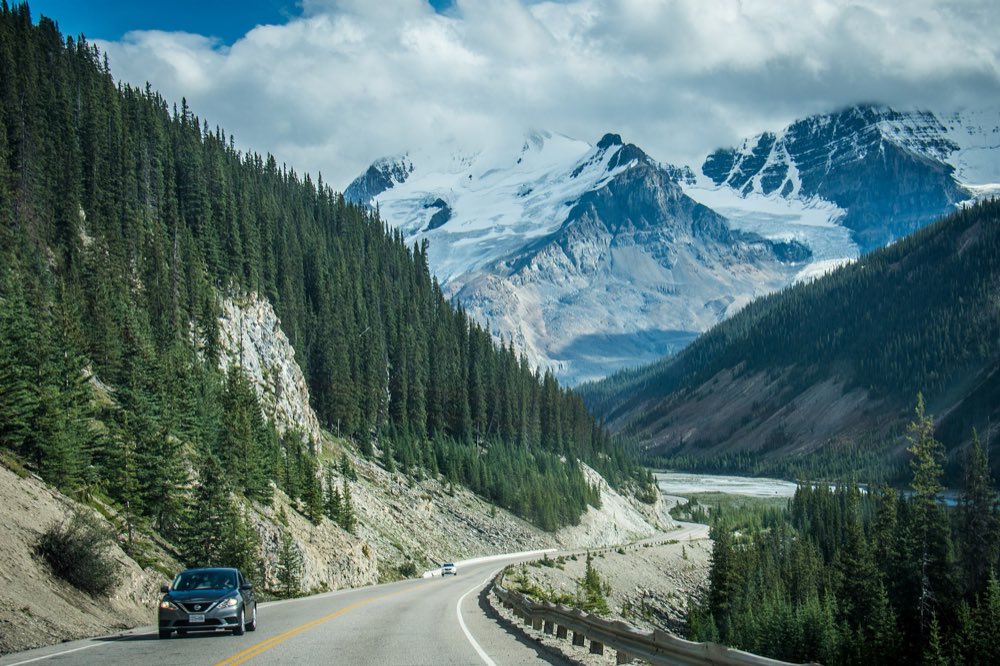 The Icefields Parkway curves through dark spruce forest beside a pale cliff, with a wide river valley and snow-covered peaks and glaciers ahead