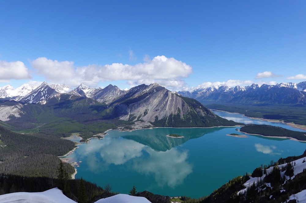 Turquoise Upper Kananaskis Lake with its forested islands spread out below grey limestone peaks and snowy ranges, seen from a high ridge with patches of snow in the foreground