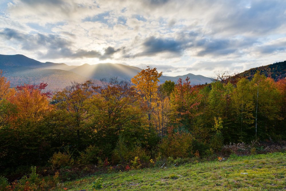 Orange, red and gold trees in late-afternoon light at a roadside overlook, with sunbeams breaking through clouds over a line of blue mountain ridges