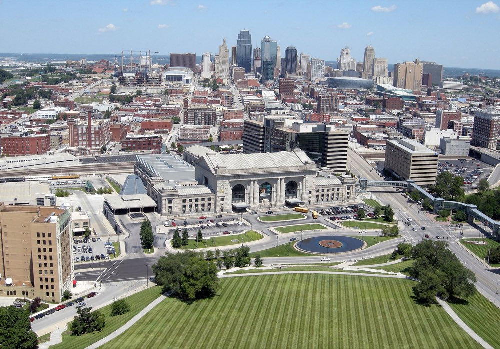 Looking north over the striped lawn below the Liberty Memorial to the long stone front of Union Station, with the downtown Kansas City skyline behind it under a blue sky