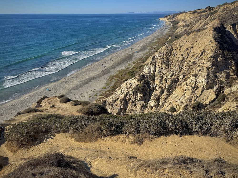 Eroded tan sandstone cliffs and coastal scrub dropping to a long empty beach, with white surf and blue ocean stretching north under a clear sky