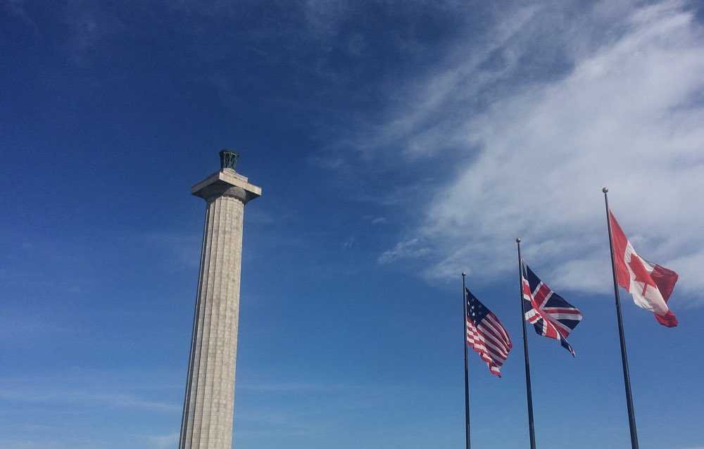The granite column of Perry's Victory memorial against a deep blue sky with wispy clouds, beside flagpoles flying the flags of the United States, the United Kingdom and Canada