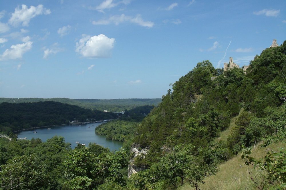 A wooded bluff above a winding green arm of Lake of the Ozarks dotted with boats, with the stone castle ruins of Ha Ha Tonka on the ridge top under a blue summer sky