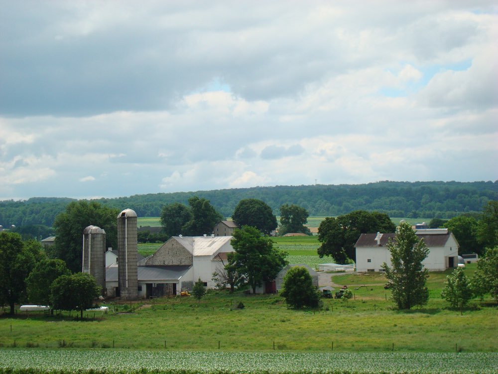 A white Lancaster County farmstead with two stone silos, barns and a farmhouse among green fields and trees, with a wooded ridge beyond under a cloudy summer sky