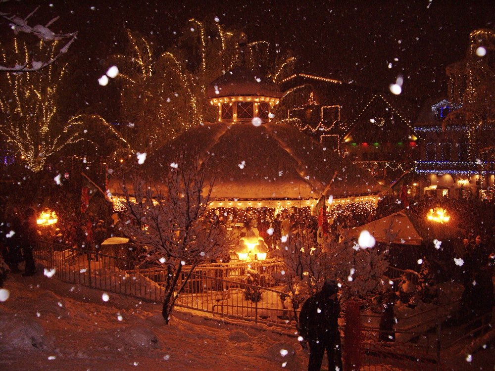 The Front Street Park gazebo in Leavenworth at night under fresh snow, with light-wrapped trees, lit Bavarian-style buildings and snow falling