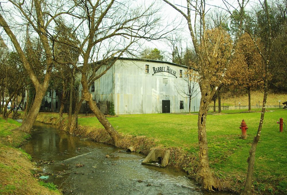 A long, pale corrugated-metal barrel house among bare trees beside a shallow creek and green lawn at the Jack Daniel Distillery in Lynchburg