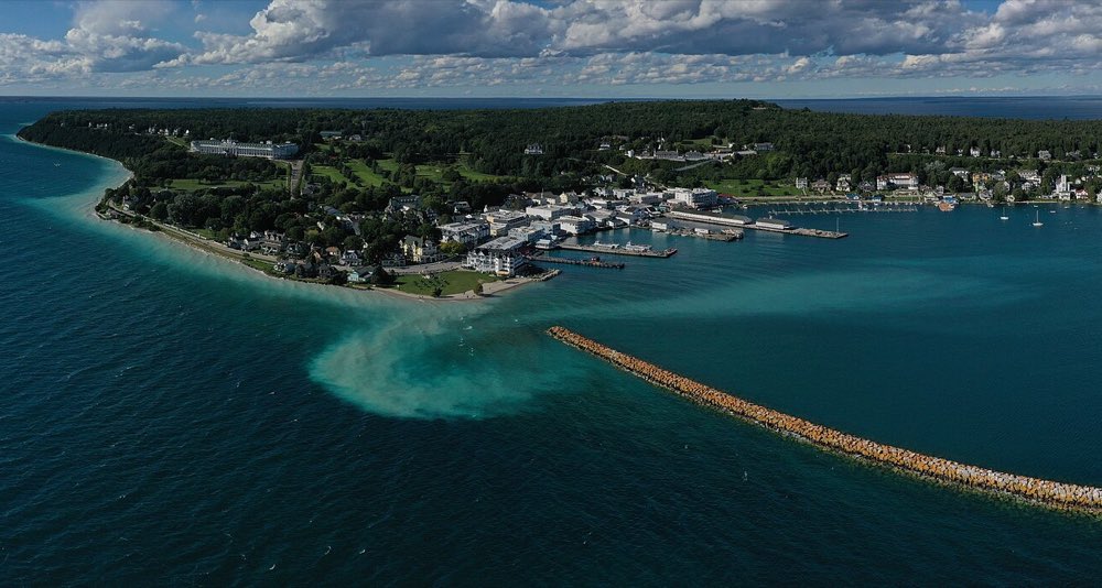 Aerial view of Mackinac Island's harbor, with white hotels, ferry docks and sailboats along the shore below a wooded bluff, a long stone breakwater and turquoise shallows in the foreground