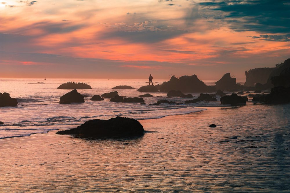 Sea stacks and boulders silhouetted on a wet beach under an orange and violet sunset sky, with surf washing in