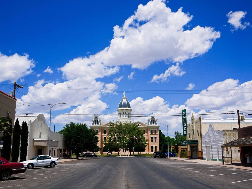 A wide, quiet street in downtown Marfa leading to a domed sandstone-colored courthouse under a deep blue sky with big white clouds, with a vintage theater sign on the right