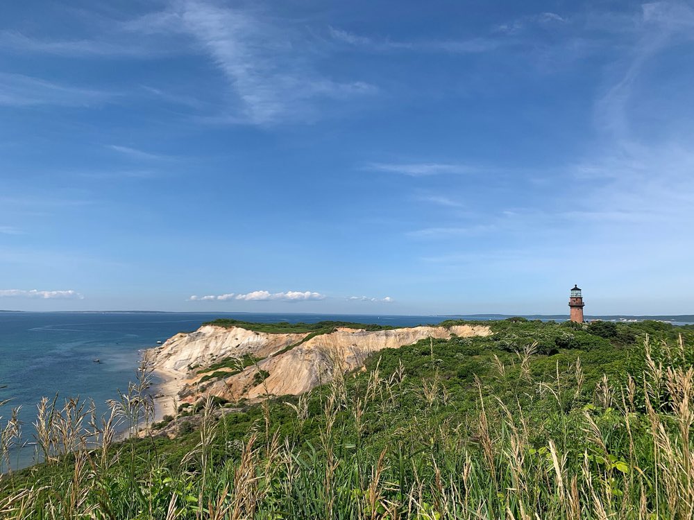 Green grassy headland above pale clay cliffs at the water's edge, with a red brick lighthouse on the far bluff and blue sea beyond