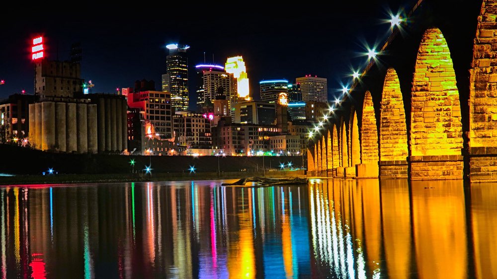 The Stone Arch Bridge lit up at night, its golden arches and the downtown Minneapolis skyline reflected in the dark Mississippi River