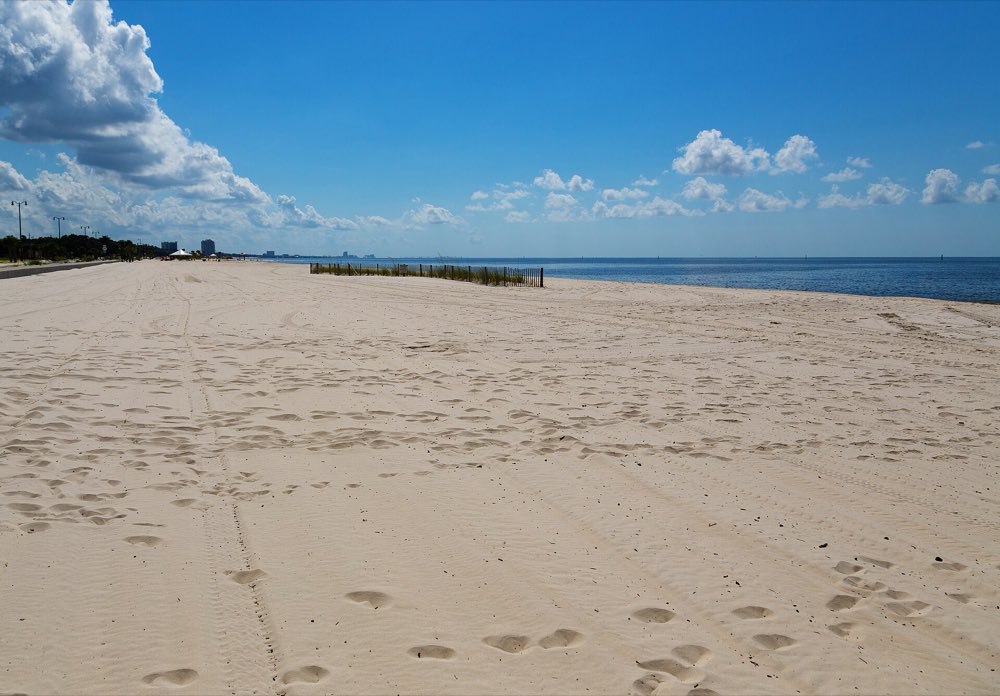 A wide, empty stretch of pale sand beach with footprints, a line of dune fencing and calm blue water of the Mississippi Sound under a bright sky