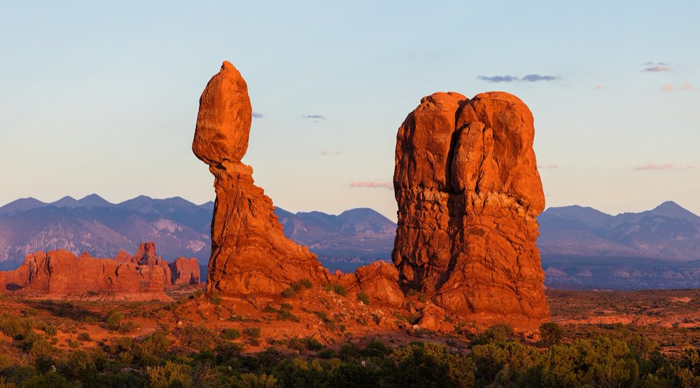Balanced Rock, a boulder perched on a tall sandstone pedestal, glowing red at sunset with the La Sal Mountains behind