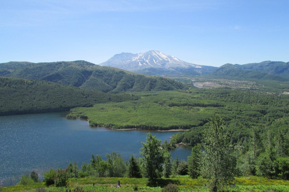 Snow-streaked Mount St. Helens rising beyond green ridges and the blue water of Coldwater Lake