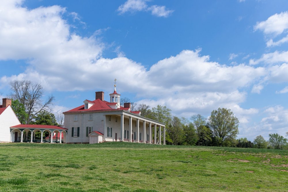 The long white mansion at Mount Vernon with its red roof, cupola and tall two-storey columned piazza, seen across a sloping green lawn under a blue sky with white clouds