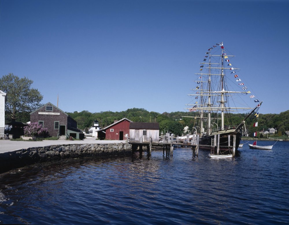 The three-masted whaleship Charles W. Morgan dressed with signal flags at a wooden pier, beside red and gray clapboard buildings on the Mystic River under a clear blue sky