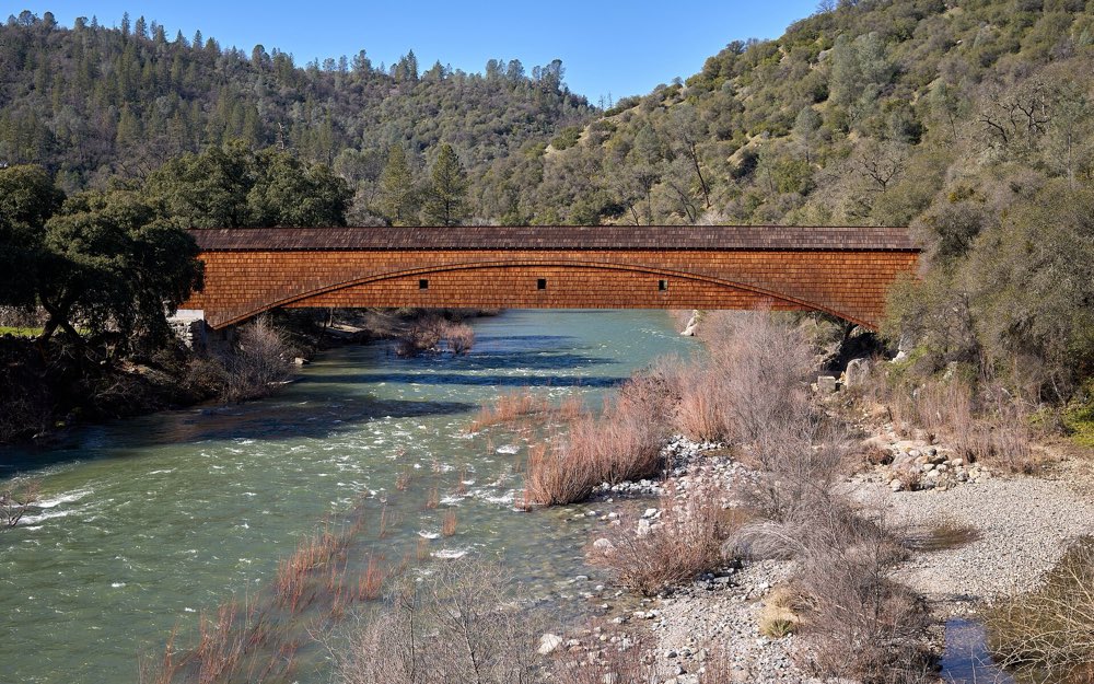 The long, shingled Bridgeport covered bridge spanning the green South Yuba River, with oak- and pine-covered hills behind