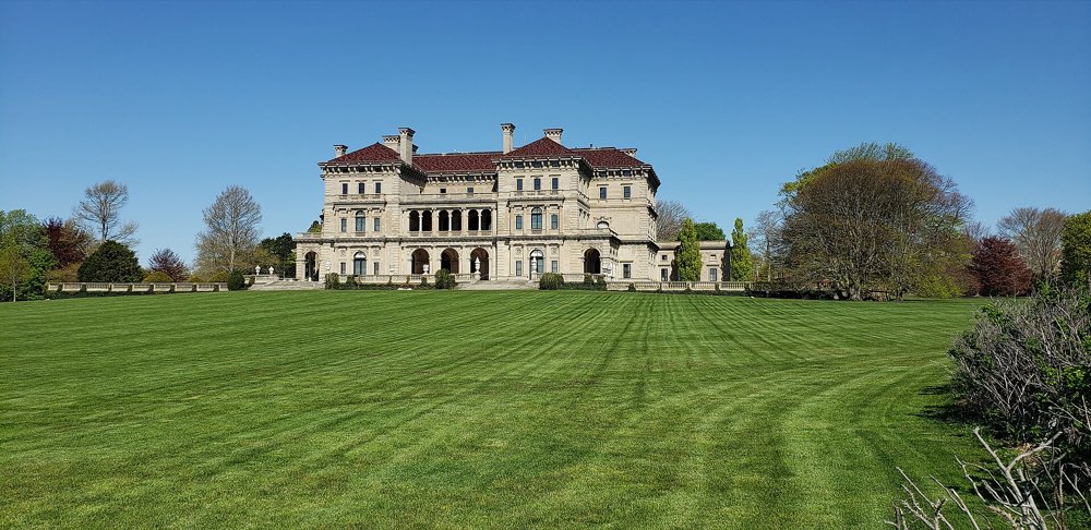 The pale stone façade of The Breakers with a red tile roof, seen across a wide striped lawn under a clear blue sky