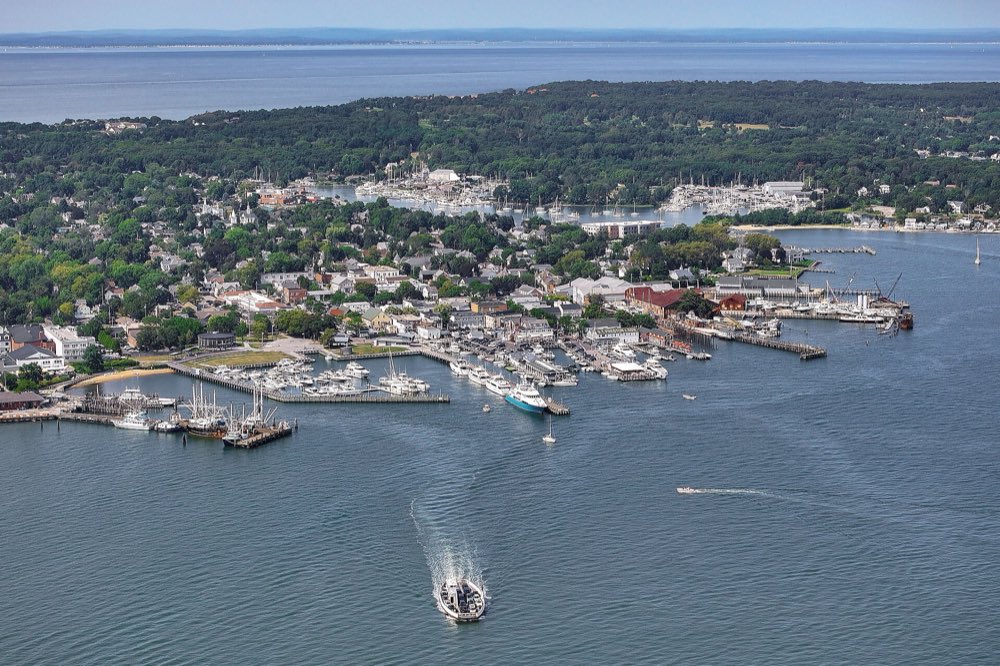 An aerial view of Greenport village on a summer day: white houses among green trees, boats tied up along the harbor docks, and a small boat leaving a wake across blue water in the foreground