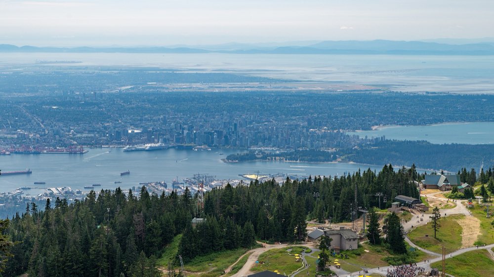 Looking down from Grouse Mountain over dark green forest to Burrard Inlet, Stanley Park and the towers of downtown Vancouver, with the flat delta and distant islands beyond