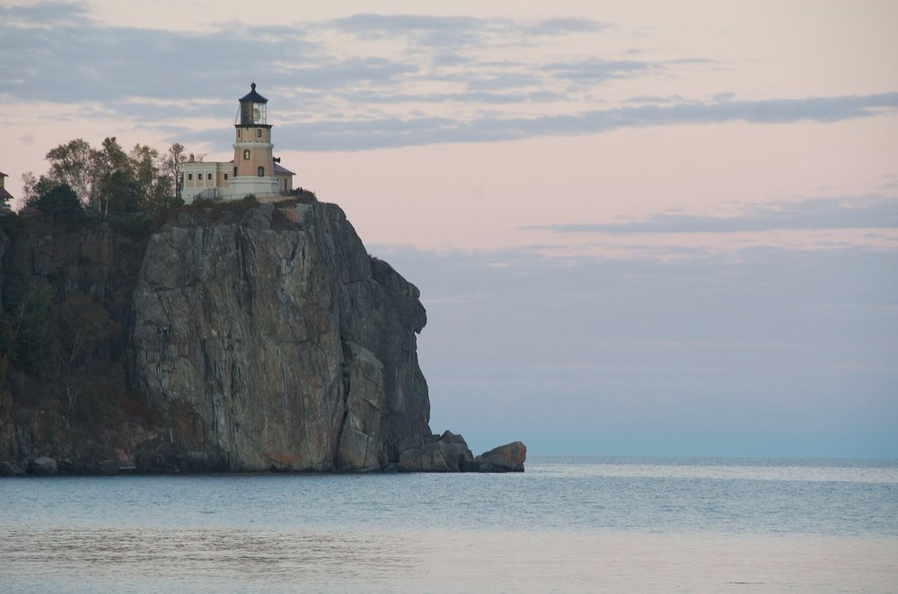 Split Rock Lighthouse on top of its sheer rock cliff above a calm Lake Superior at dawn, with pale pink clouds behind