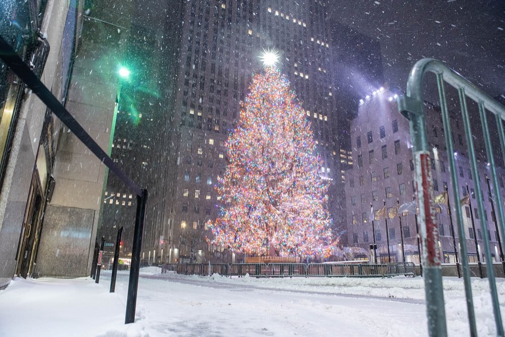 The Rockefeller Center Christmas tree glowing with multicolored lights in falling snow, seen from a snowy, empty plaza with tall stone towers behind