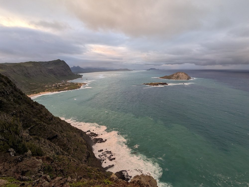 View from the top of Makapuʻu Point down a dark green sea cliff to a turquoise bay, a curve of white sand and the domed island of Mānana offshore under a cloudy sky