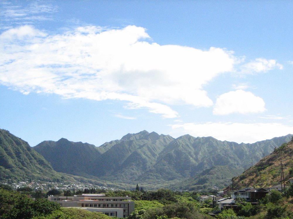 Green, steep-sided ridges of the Koʻolau Range closing in around the head of Mānoa Valley under white clouds, with rooftops and trees in the foreground