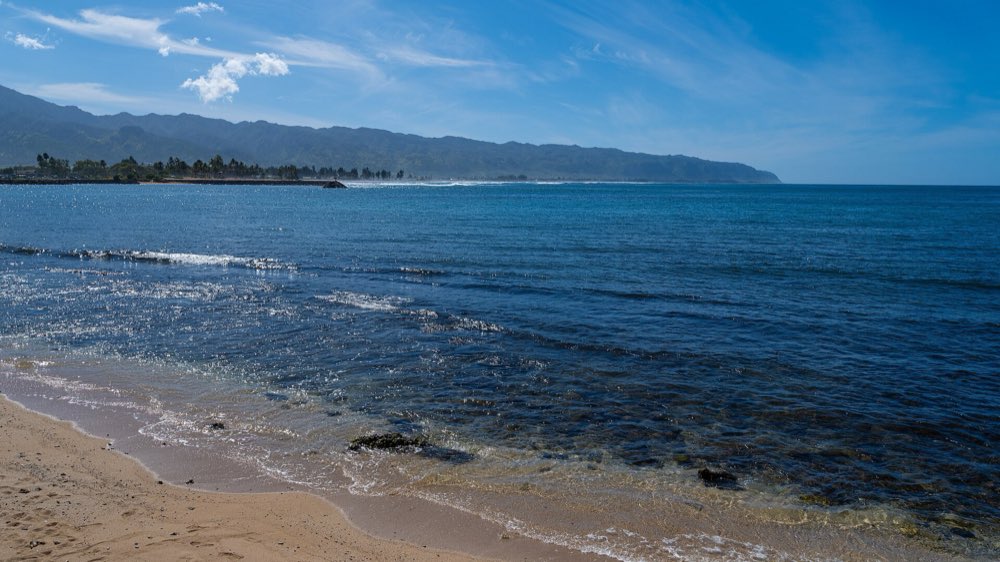 Clear blue water lapping a sandy beach at Haleʻiwa, looking across the bay to a breakwater, a line of trees and the long green ridge of the Waiʻanae Range under a light-clouded sky