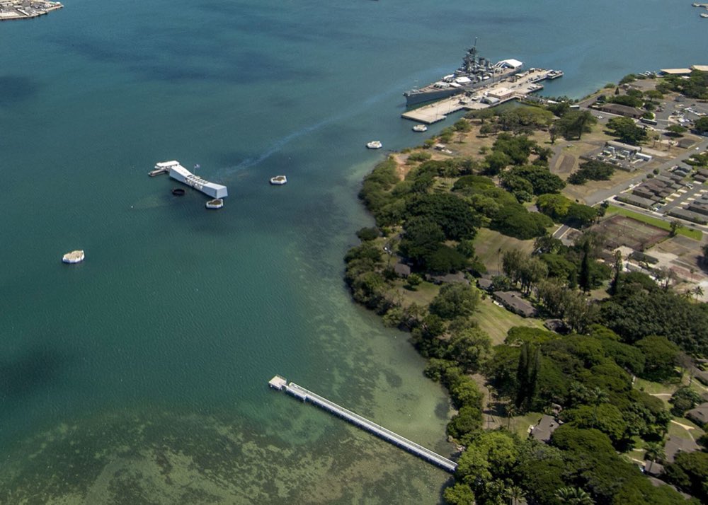 Aerial view over the turquoise water of Pearl Harbor: the white USS Arizona Memorial on the left, the grey Battleship Missouri moored at a pier on the right, and the green shore of Ford Island between them