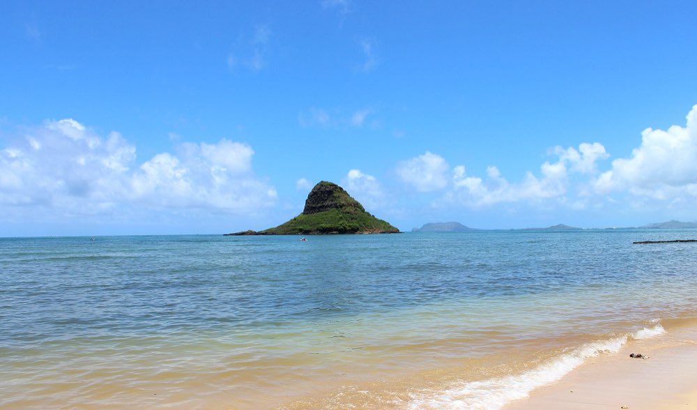 The steep, tree-topped islet of Mokoliʻi rising from calm blue-green water off a sandy shore, with low islands on the horizon under scattered white clouds
