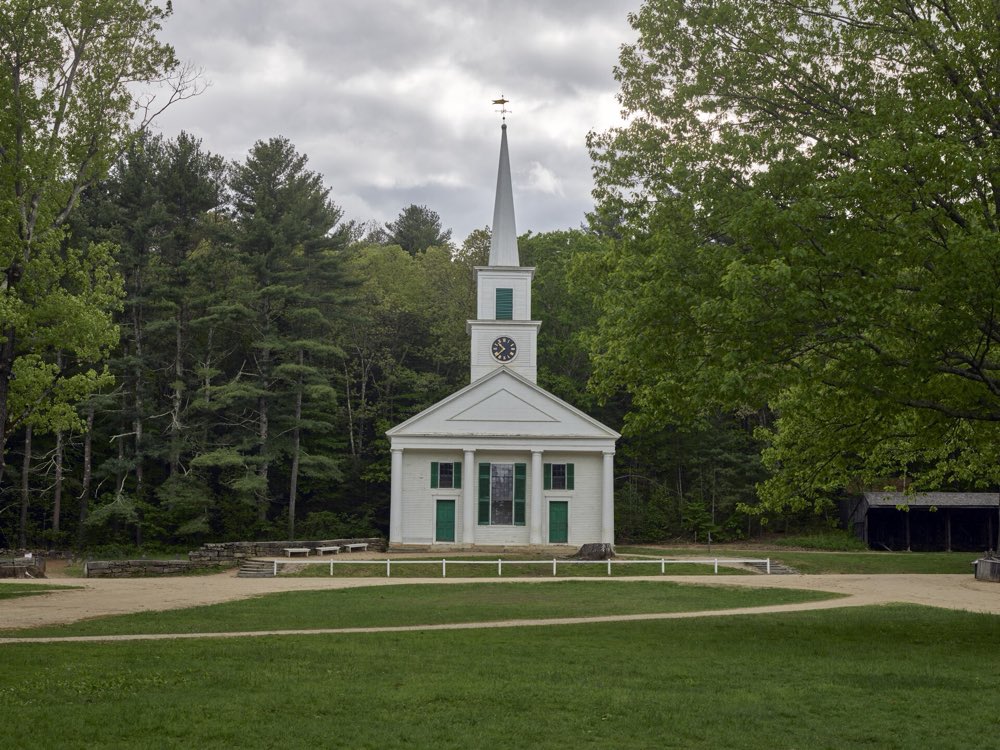 A white meetinghouse with green shutters, a clock tower and a tall spire at the end of a grassy common, with dirt paths and spring woods behind
