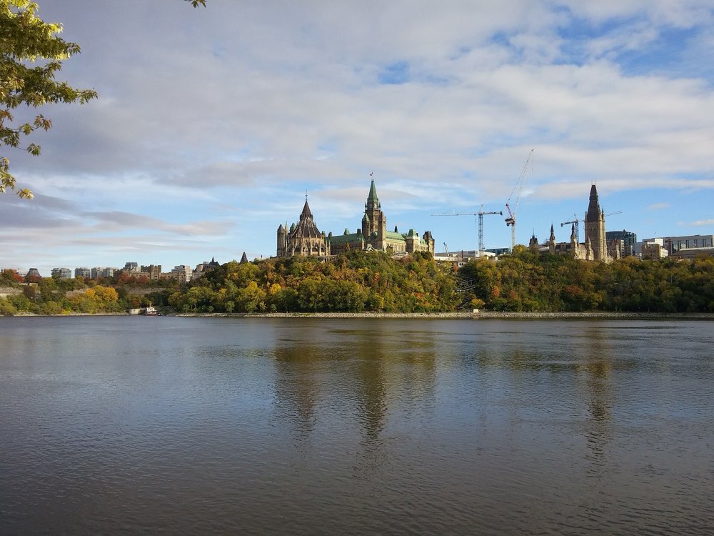 The Gothic towers of Parliament Hill rise above autumn trees on a cliff over the wide, calm Ottawa River under a blue sky with white clouds