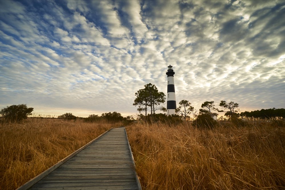 Bodie Island Lighthouse at the end of a wooden boardwalk through golden marsh grass under a rippled evening sky