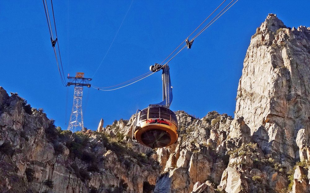 A round red-and-silver tram car hanging from cables in front of jagged granite cliffs under a deep blue sky, with a steel tower behind