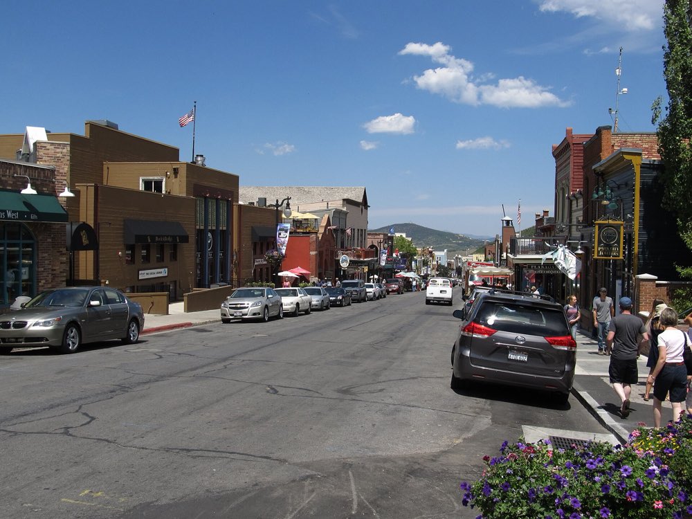 Park City's Main Street on a sunny summer day, with brick and timber storefronts, parked cars and green hills at the far end of the street