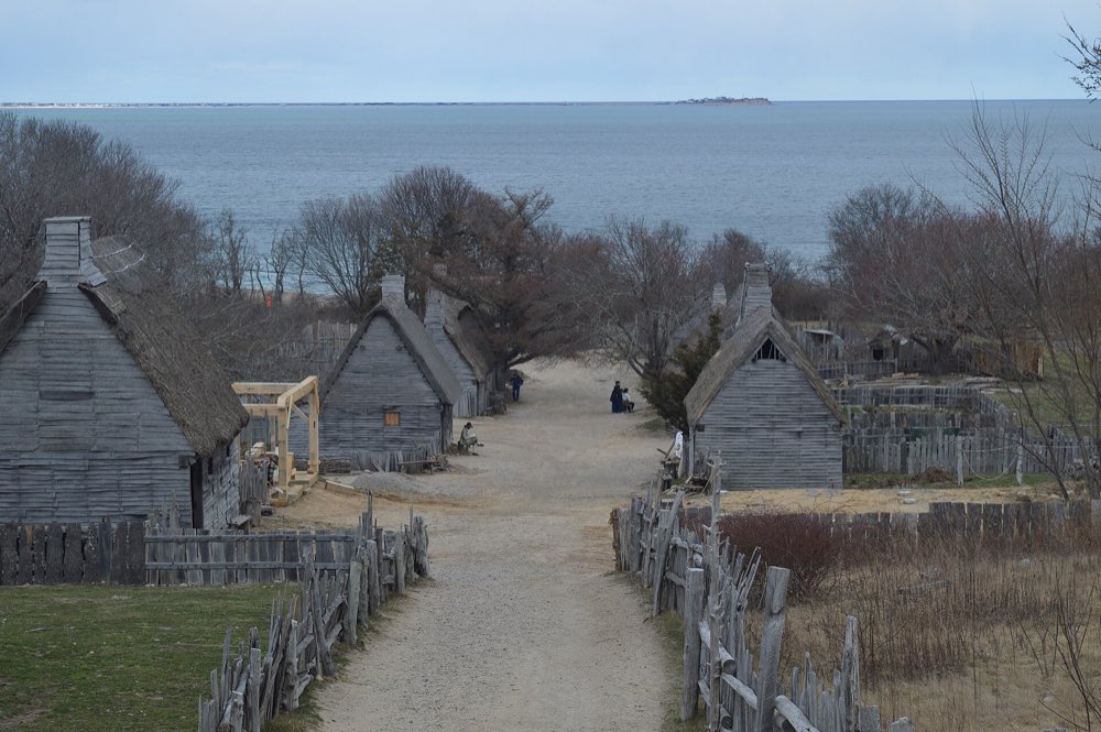 A sandy lane runs downhill between gray clapboard houses with thatched roofs and split-rail fences toward the open water of Cape Cod Bay under an overcast sky