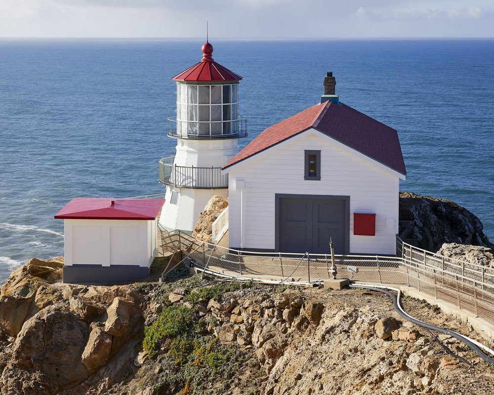 The white Point Reyes Lighthouse with its red roof and lantern room on a rocky ledge above a calm blue Pacific, the fog-signal building beside it