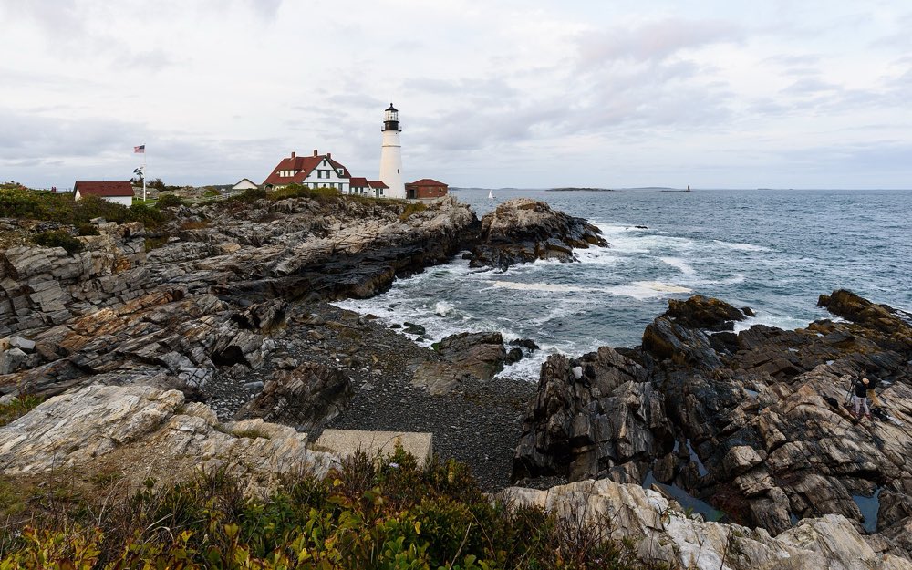 Portland Head Light, a white tower beside a keepers' house with a red roof, on a rocky point with waves breaking in a cove below under a gray sky