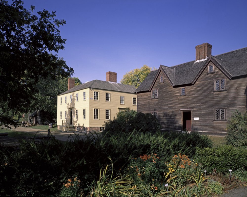A pale yellow Georgian house and a dark wooden colonial house with diamond-pane windows behind a summer garden bed under a deep blue sky