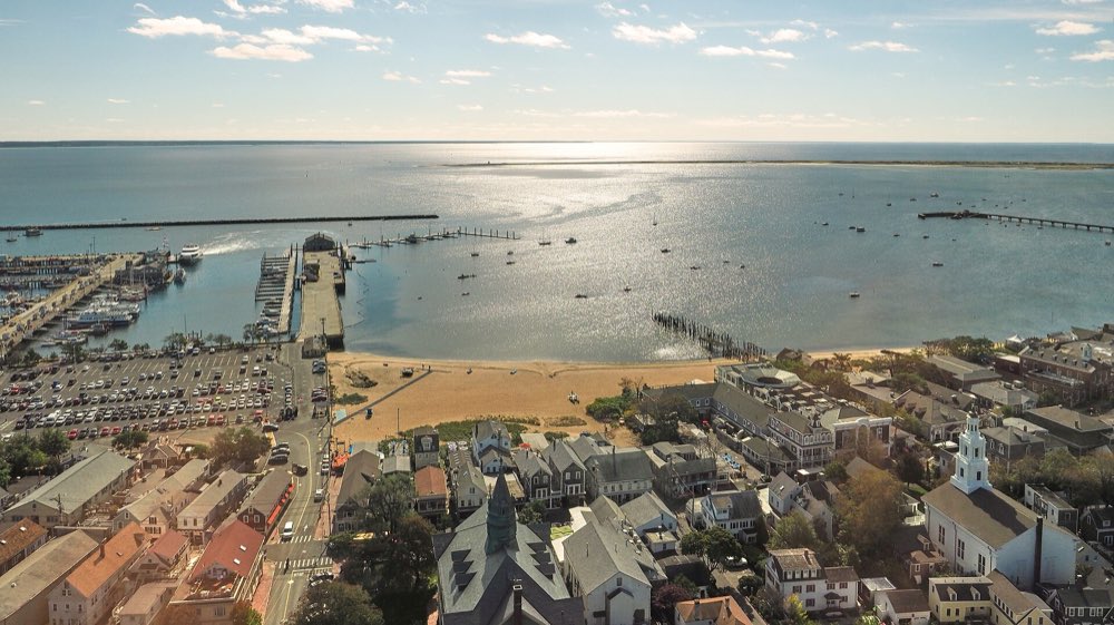 Provincetown seen from high above: rooftops and a white church steeple in front, a curving sandy harbor beach, MacMillan Pier reaching into calm blue water and the long breakwater beyond