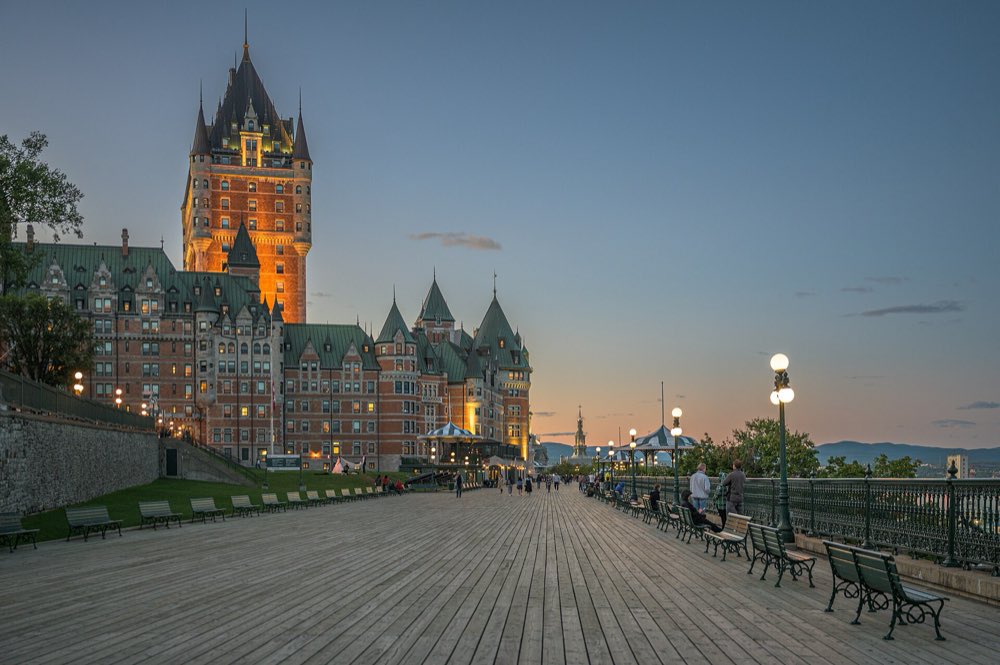 The Château Frontenac's copper roofs and brick tower glowing at dusk above the long wooden boardwalk of Terrasse Dufferin, lined with benches and lamp posts beside the river