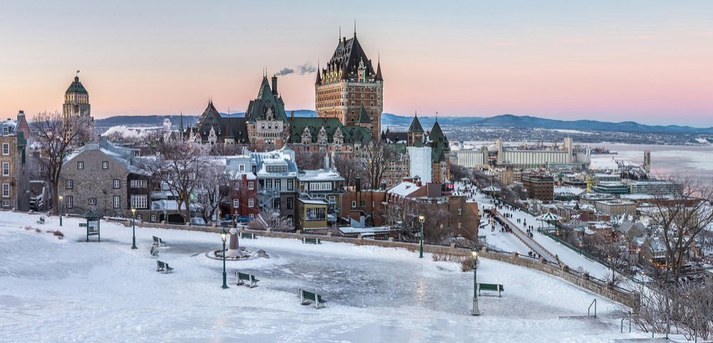 The Château Frontenac and the rooftops of Old Québec glazed with ice and snow at dusk, with a frozen park and lamp posts in front and the white St. Lawrence beyond