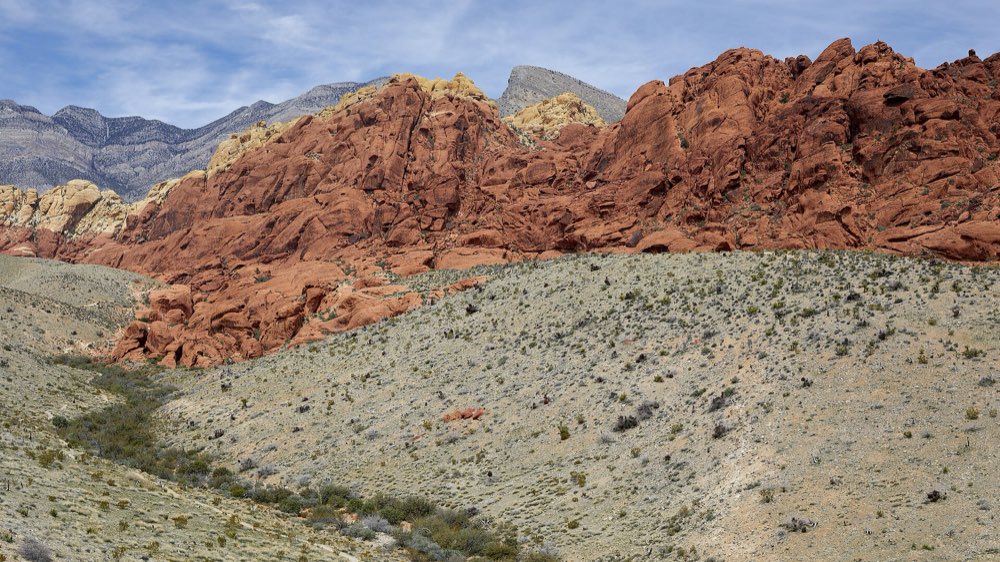 Rounded red and cream sandstone of the Calico Hills rising above a pale desert slope dotted with low shrubs, with grey limestone peaks behind under a lightly clouded sky